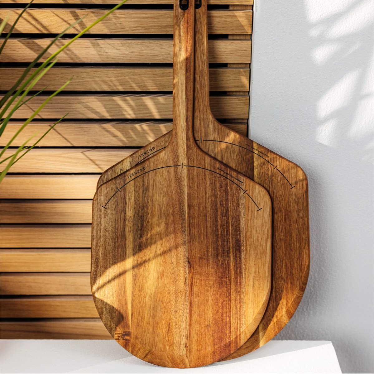 Two natural acacia wood pizza peels, one large and one standard, with etched guides and metal hanging loops.