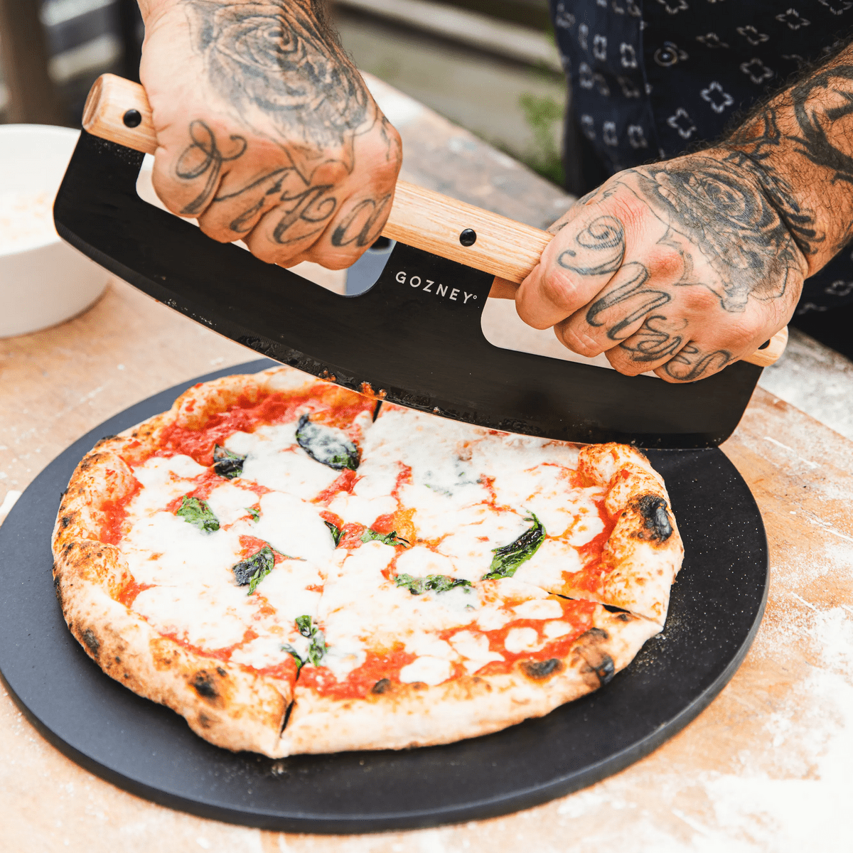 Tattooed hands using a black Gozney pizza server with a light wood handle to slice a Margherita pizza on a black board.