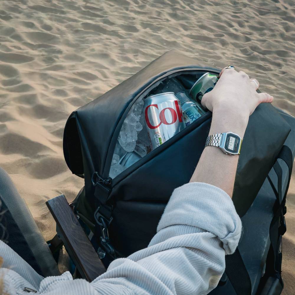 Black TentBox Cool Bag on sand, open with ice and drinks, a hand reaching inside.
