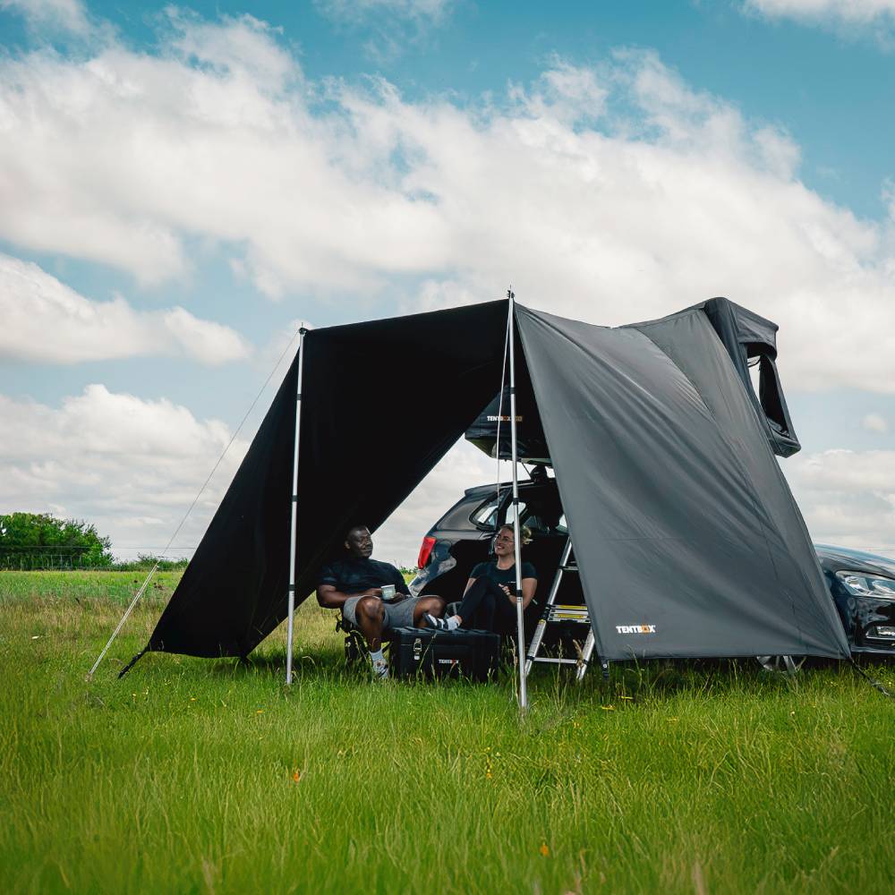 Black TentBox Go Tunnel Awning attached to a car, providing shade for two people in a grassy field.
