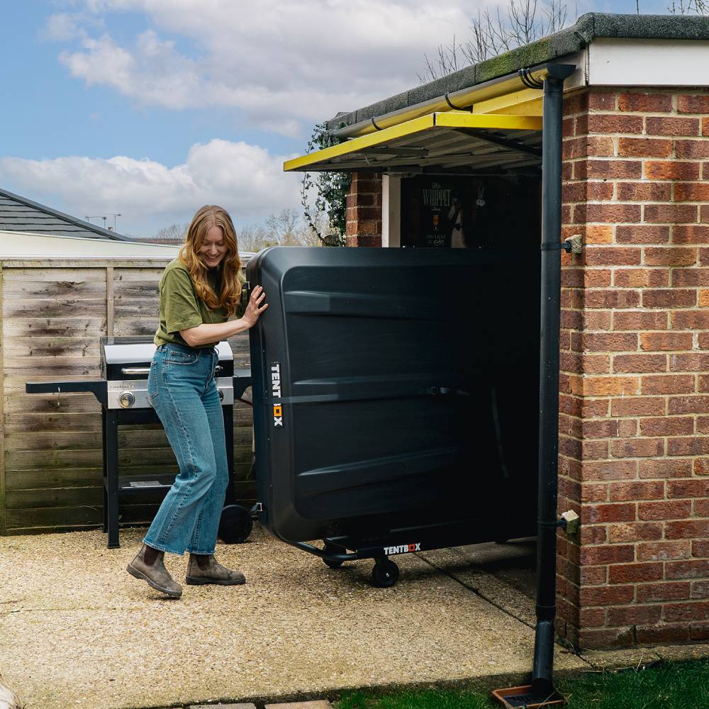 Woman pushing a large, black TentBox storage trolley with wheels into a brick garage.