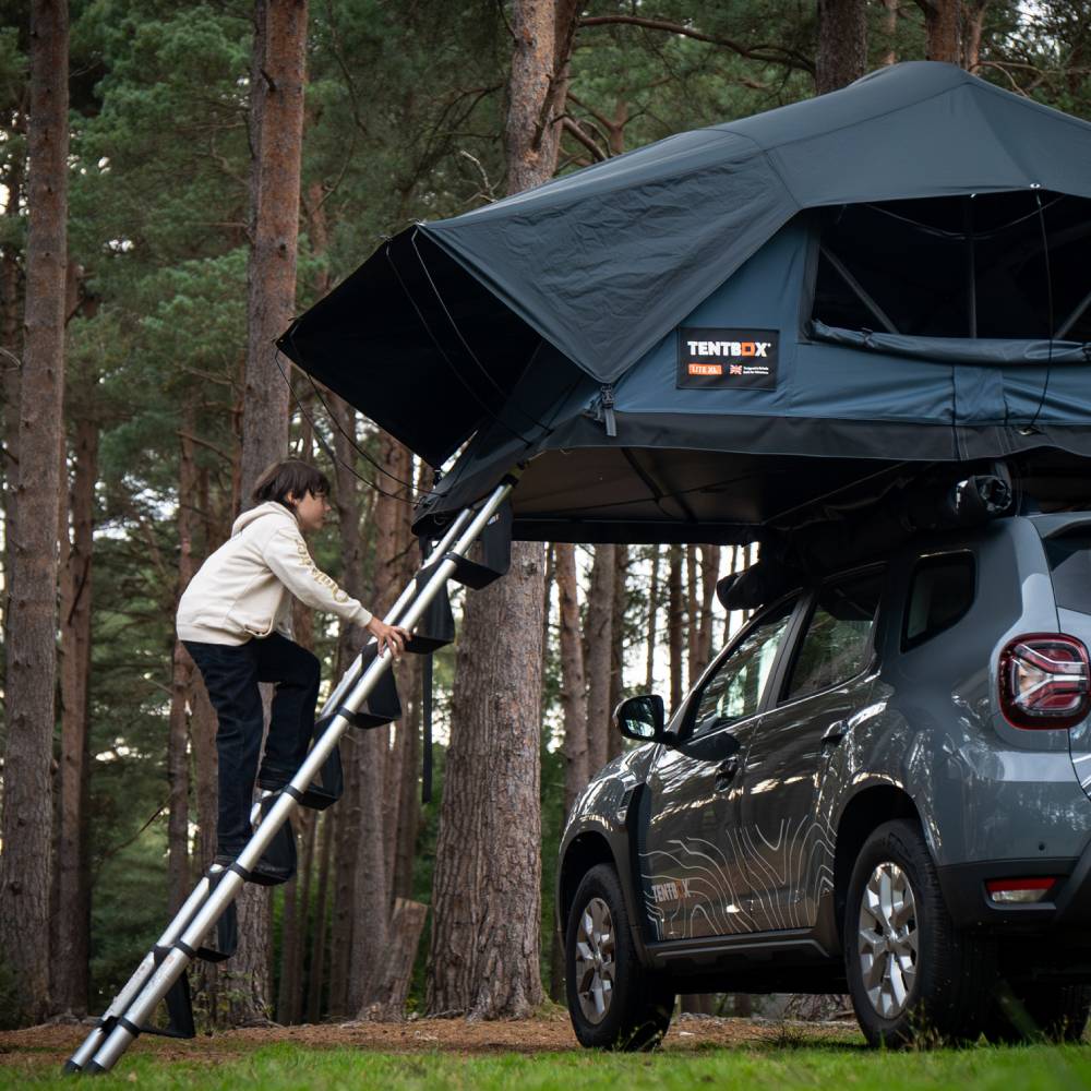 Child climbing a silver TentBox ladder with wide, anti-slip steps to a dark grey rooftop tent on a grey SUV.
