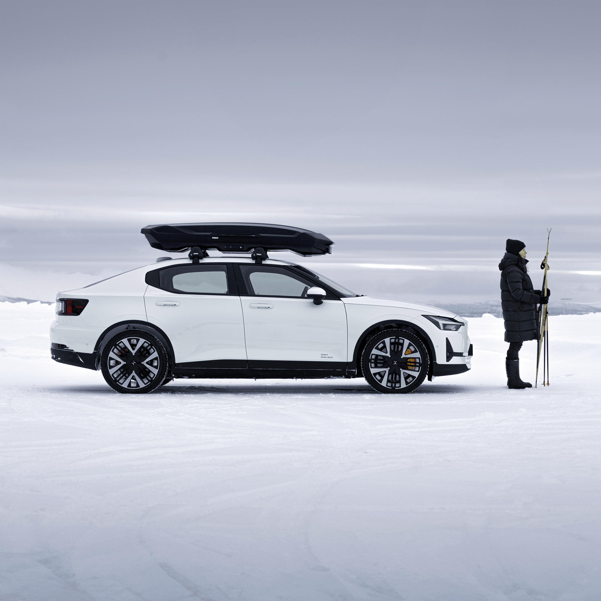 White car with a glossy black Thule Motion 3 L roof box on a snowy landscape, person with skis.