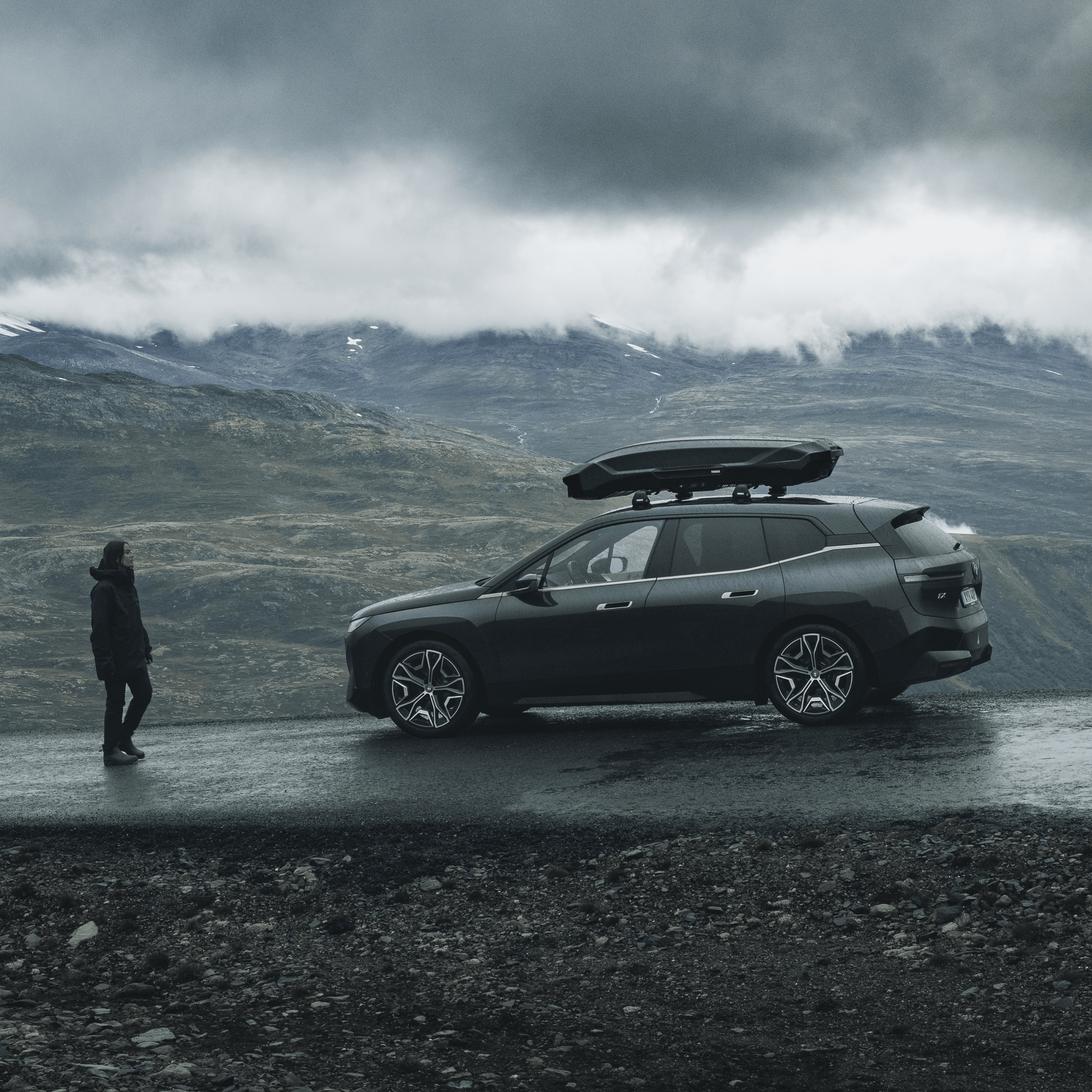 Black glossy Thule Motion 3 L roof box on a dark SUV in a cloudy mountain landscape.