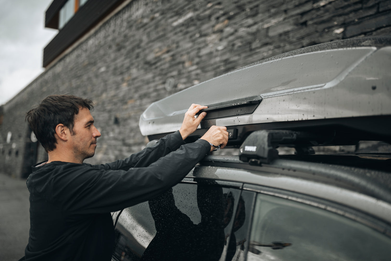 Man in black shirt attaching a dark grey Thule roof box to a car's roof rack.