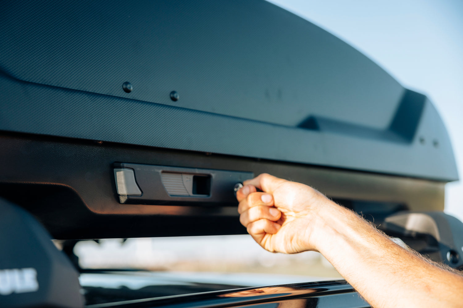Hand locking a black textured Thule roof box onto a car's roof rack.