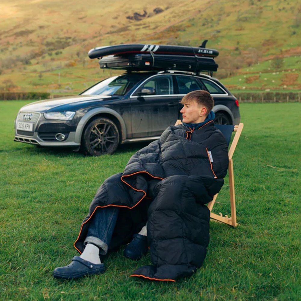 Man wrapped in a black synthetic down blanket with orange trim, sitting in a camp chair outdoors.