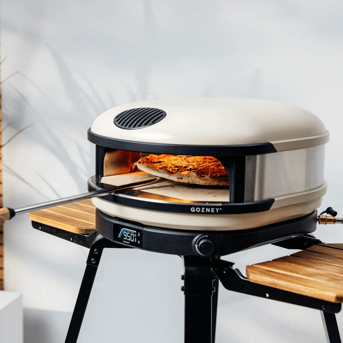 Cream Gozney Arc pizza oven on a black steel stand with acacia wood wings, cooking a pizza.