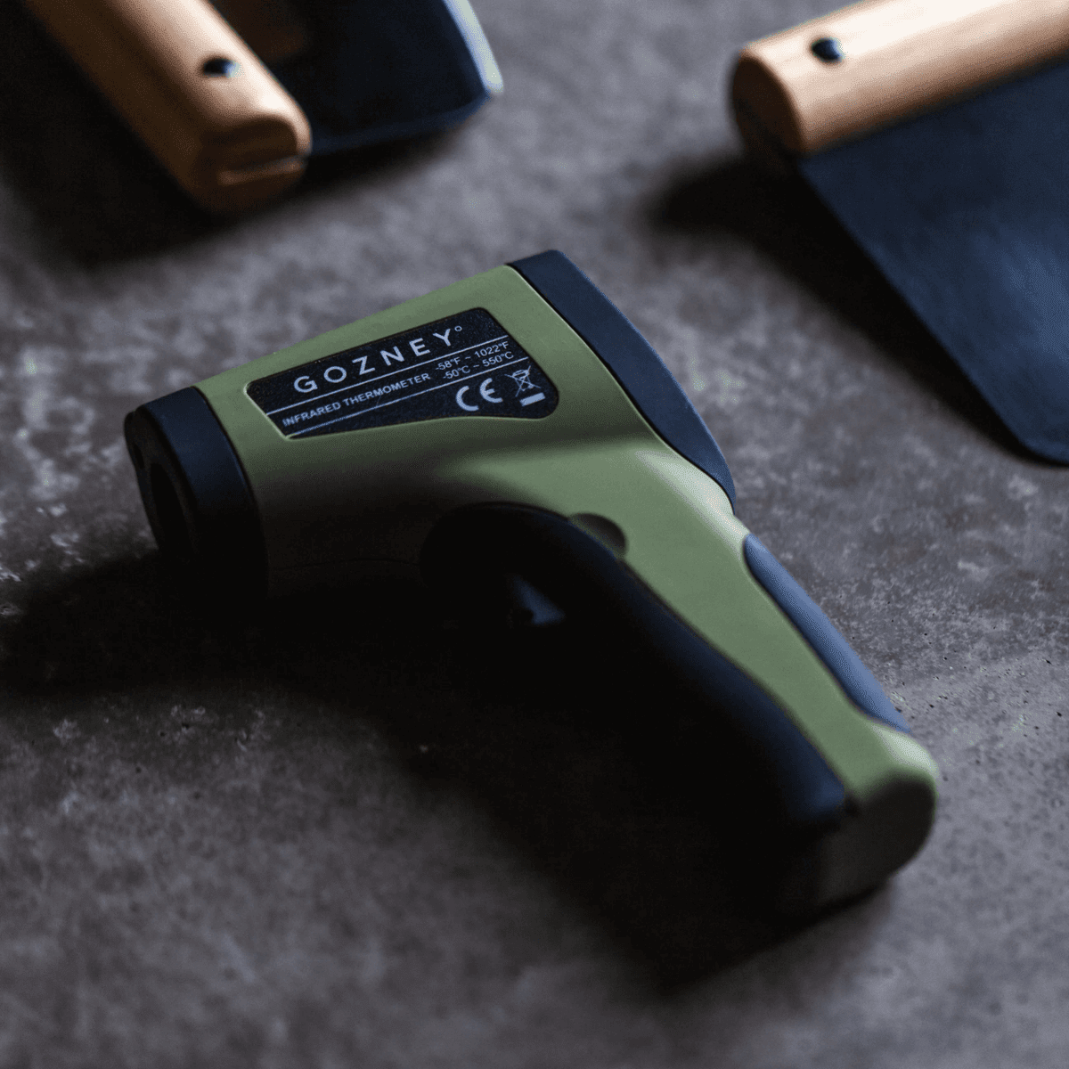 Gozney Infrared Thermometer: olive green and black, soft-touch grip, laser-guided, on a dark surface.
