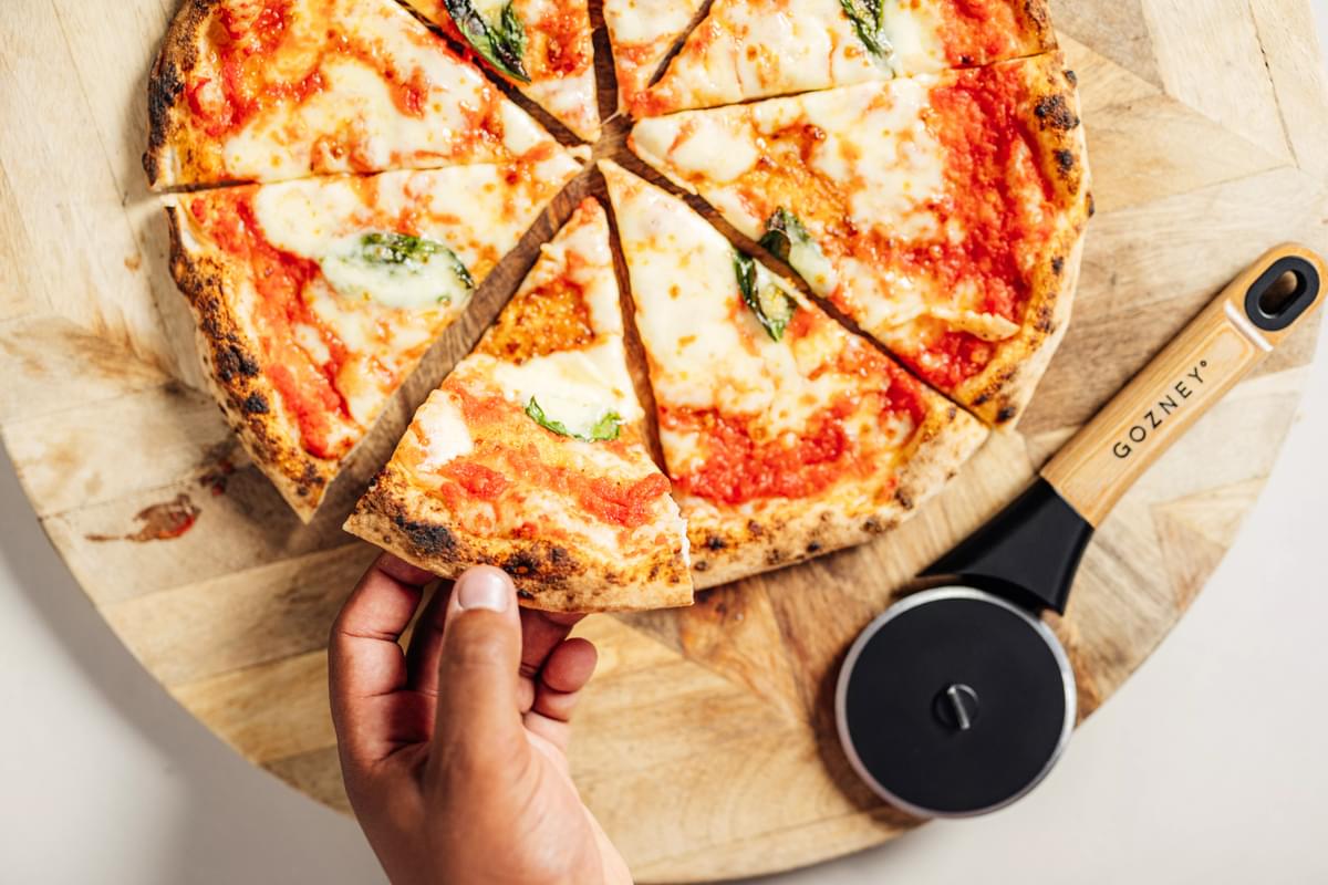 Gozney Pizza Cutter with black wheel and wood handle next to a sliced margherita pizza on a wooden board.