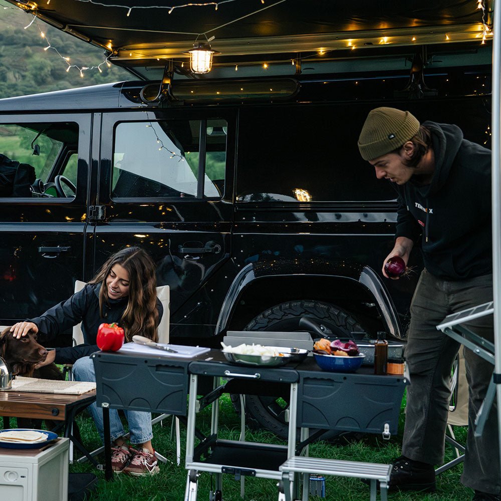 Couple cooking outdoors with a black Cargo Ultimate Bundle setup, featuring a KitchenBox and ChuckBox next to a black SUV.