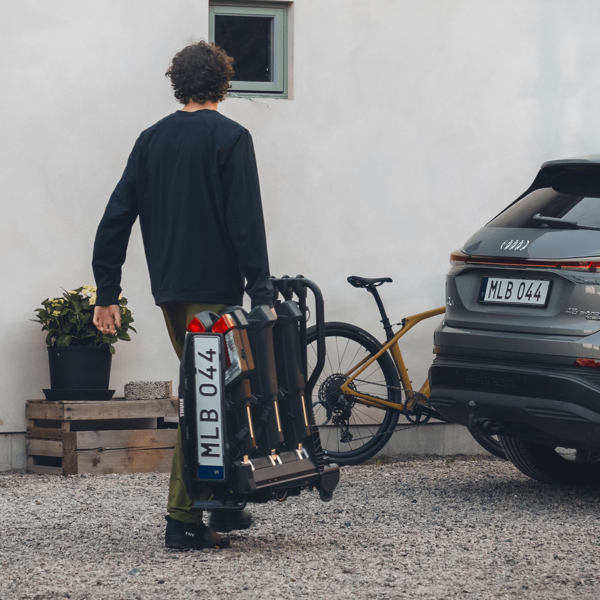 Man carrying a folded Thule EasyFold 3 bike carrier with integrated lights and license plate holder, next to a grey car and a bicycle.