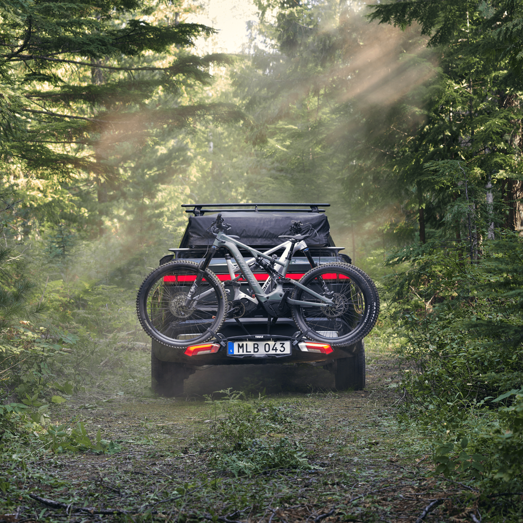 Dark grey Thule Epos 2-bike rack on a black SUV, carrying two mountain bikes on a forest trail.