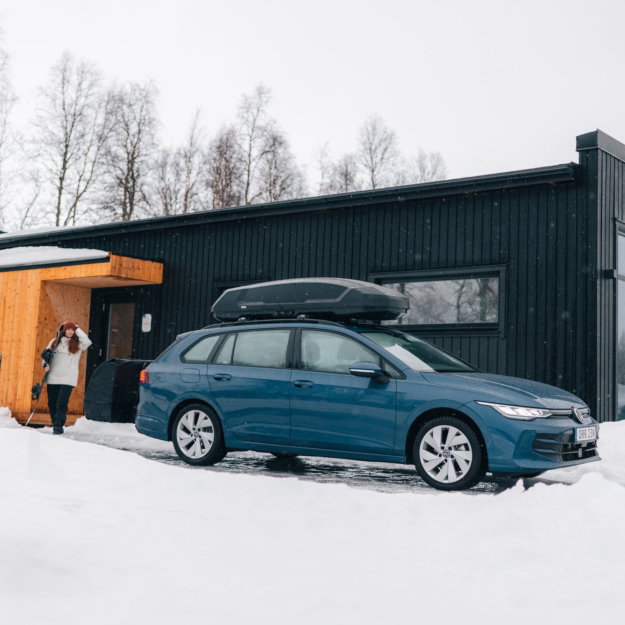 Blue car with a sleek black Thule Force 3 L Sport roof box parked in snow next to a modern house.