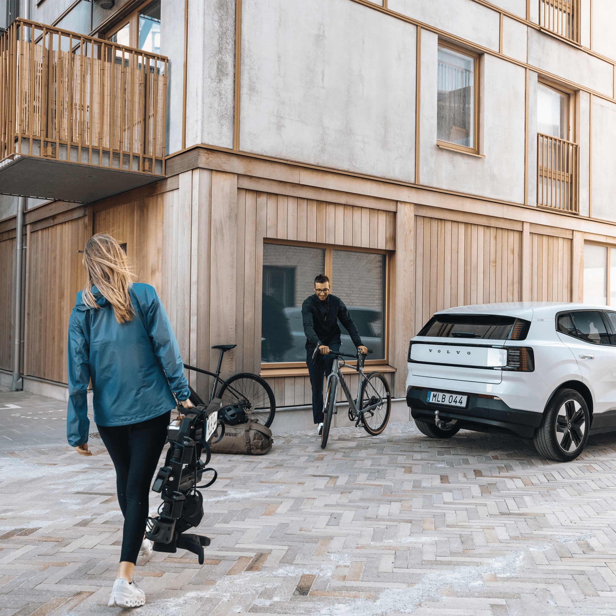 Woman carrying a black Thule OutPace 3-bike rack, man with a bicycle, and a white Volvo car.