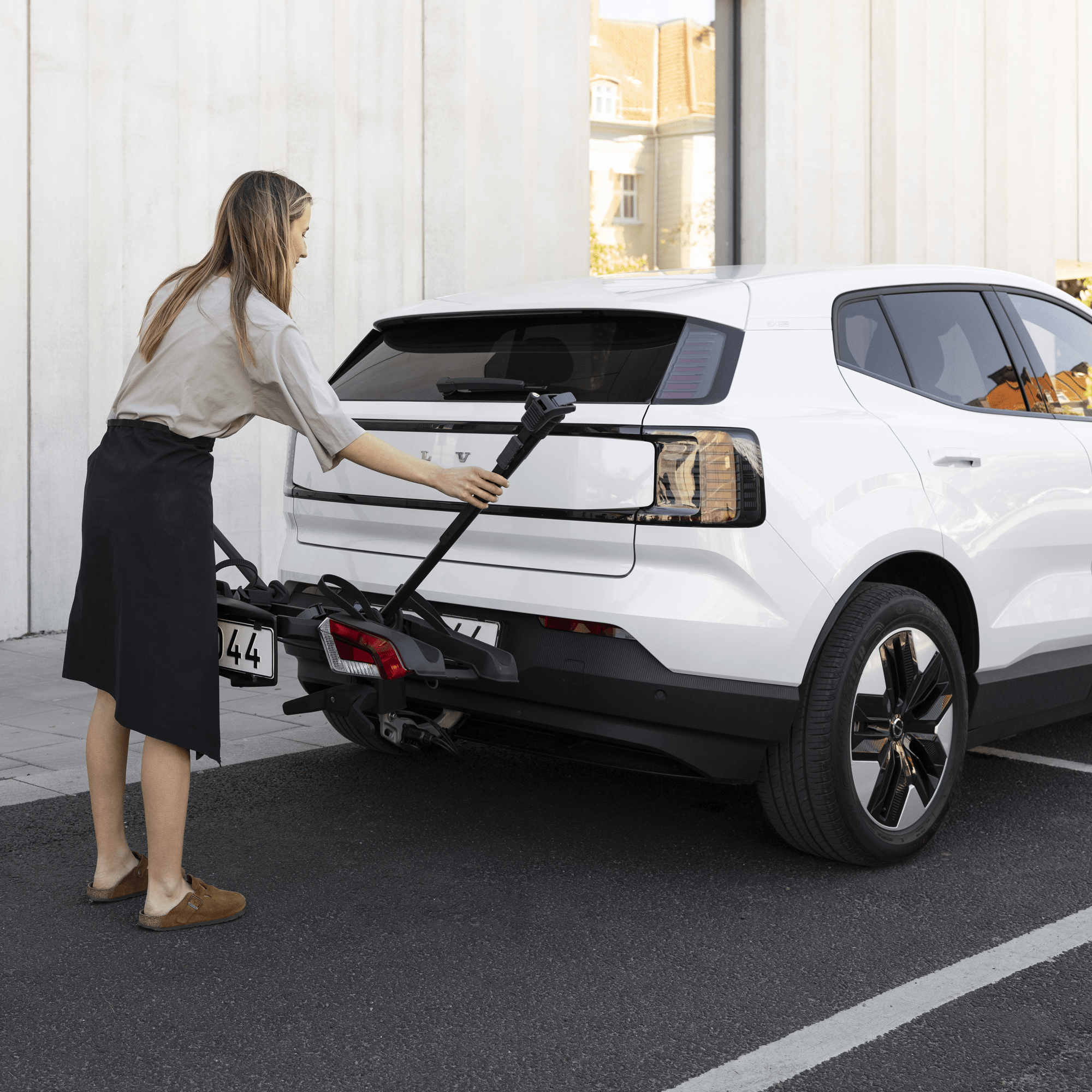 Woman attaching a black Thule OutPace 3-bike rack to the rear of a white Volvo car.
