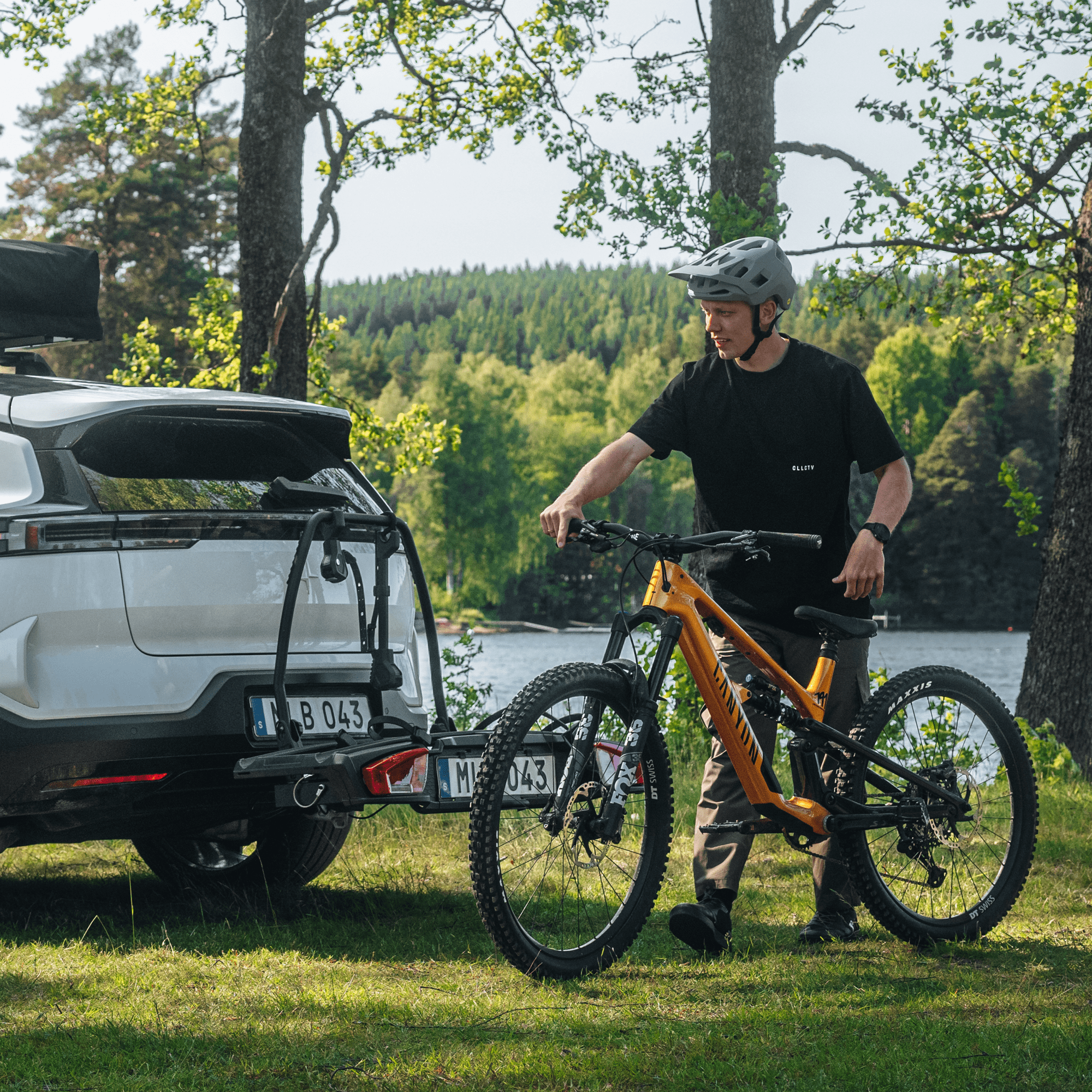 Man in helmet loading an orange mountain bike onto a black Thule VeloSpace 3 towbar bike rack on a white SUV.