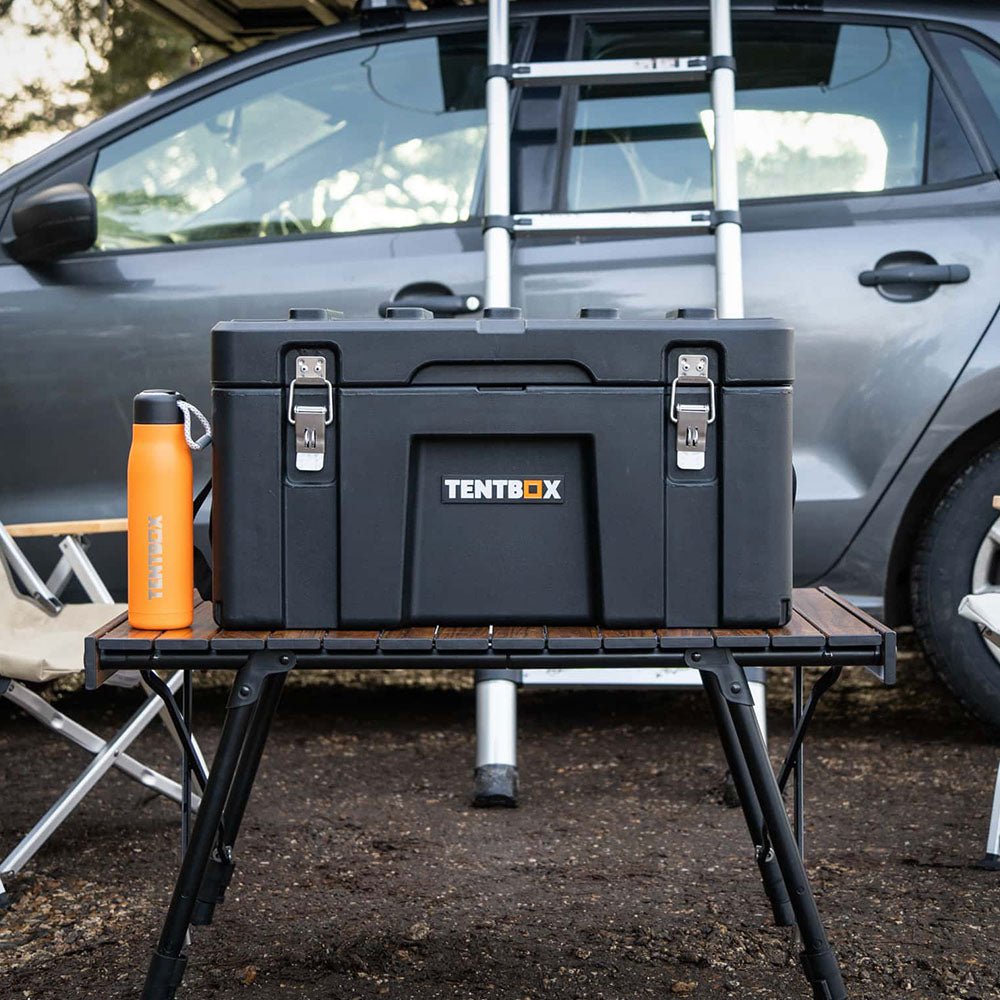 Black rugged TentBox Chuck Box with steel latches on a wooden camping table, next to an orange water bottle.