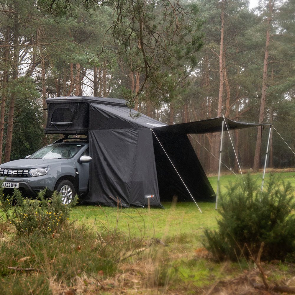 Gray SUV with a black Classic 2.0 rooftop tent and attached awning in a forest setting.