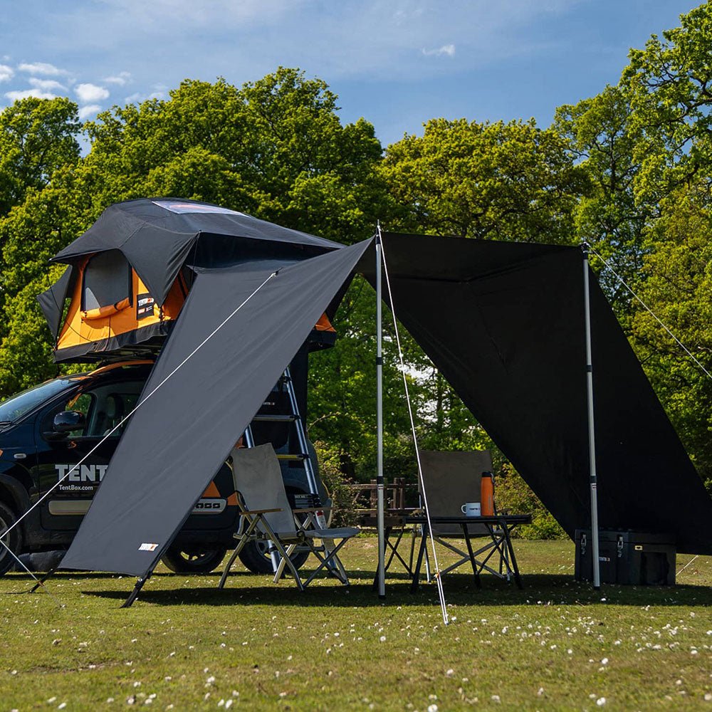 Black Lite Tunnel Awning attached to a TentBox Lite 2.0 roof tent on a car, providing shade over camping chairs and a table in a grassy field.
