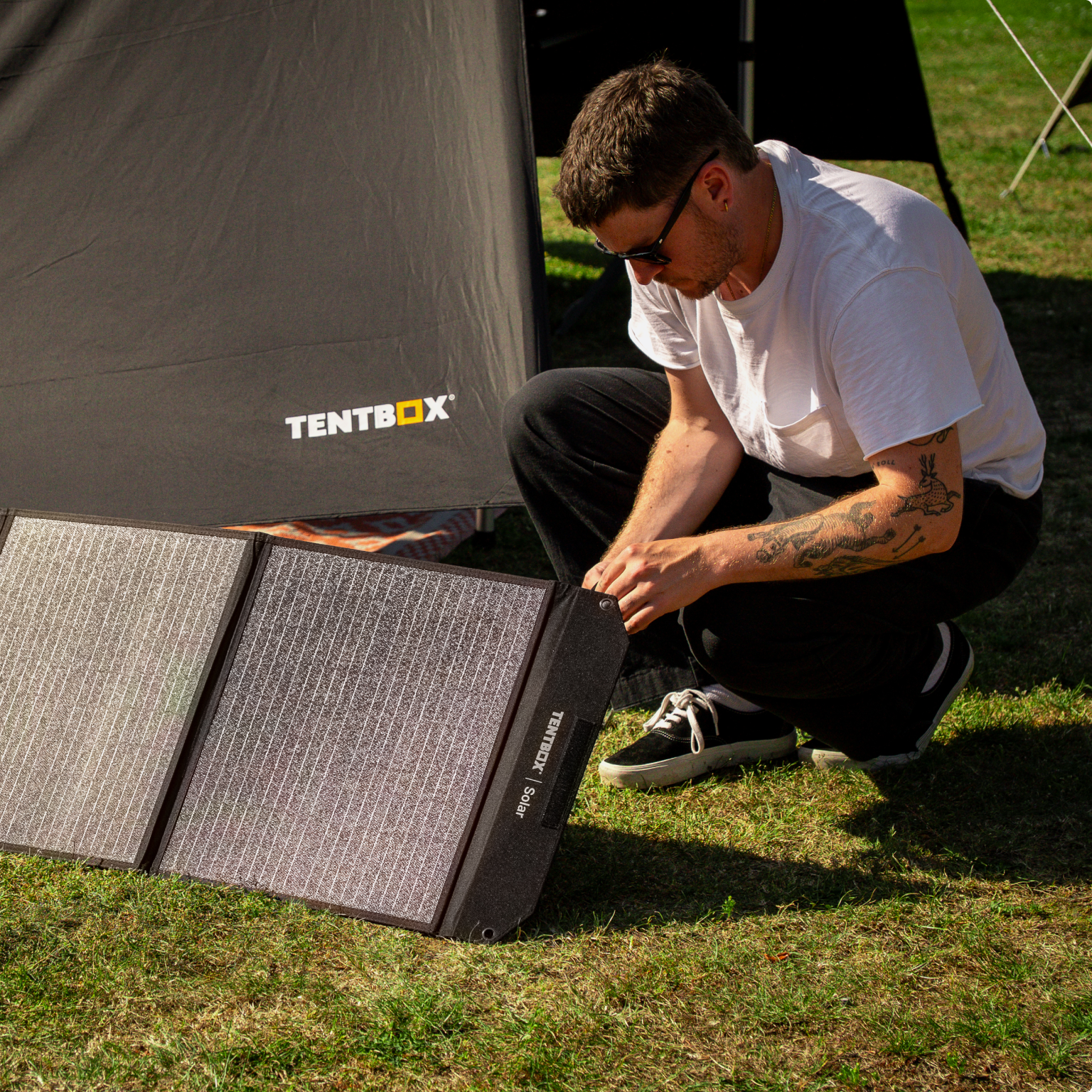 A man in a white shirt in front of a TentBox tent plugging the TentBox solar panel into a power station